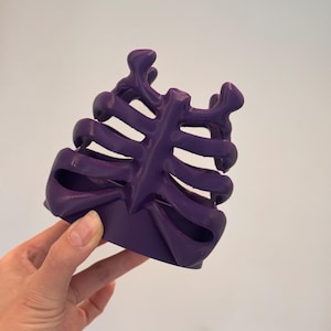 May include: A deep purple, three-dimensional object shaped like a human rib cage. The structure features a central vertical sternum piece with curved, horizontal ribs extending outward on both sides, creating an open, cage-like design.