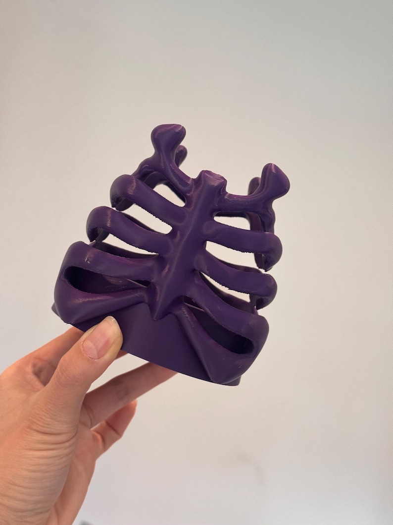 May include: A deep purple, three-dimensional object shaped like a human rib cage. The structure features a central vertical sternum piece with curved, horizontal ribs extending outward on both sides, creating an open, cage-like design.