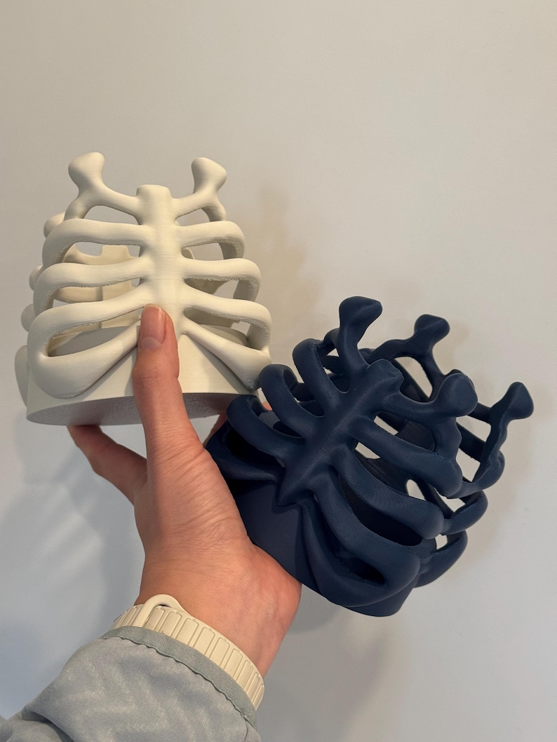 May include: Two cylindrical containers featuring a three-dimensional rib cage design. One container is matte white and the other is matte dark blue. Both items have an open-frame structure with curved, bone-like ribs extending from a central spine.