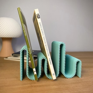 May include: A teal corrugated paper bookend holding two books. One book is green with the title "Glaxo" and the other is beige with the title "Paul Gauguin, Noa Noa, Viaje de Tahiti". The bookend has a wavy design.