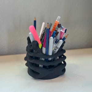 May include: A matte black pencil holder shaped like a human rib cage, filled with a variety of colorful pens, pencils, and markers. The cylindrical container features curved, bone-like structures that form the exterior walls, holding writing tools.