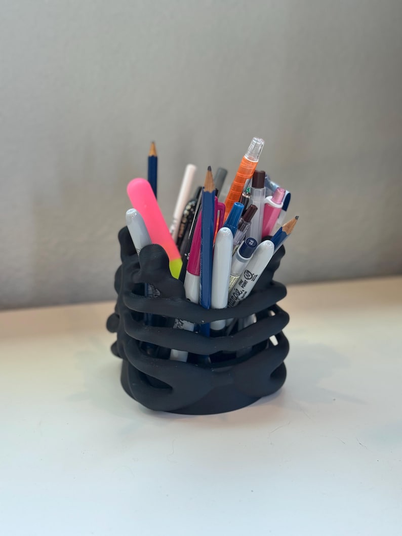 May include: A matte black pencil holder shaped like a human rib cage, filled with a variety of colorful pens, pencils, and markers. The cylindrical container features curved, bone-like structures that form the exterior walls, holding writing tools.