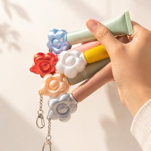 May include: A hand holding several colorful lip balm tubes. Each tube has a flower-shaped cap in shades of blue, red, white, orange, and silver. The tubes are attached to keychains. The lip balm tubes are in shades of pink, yellow, and green.