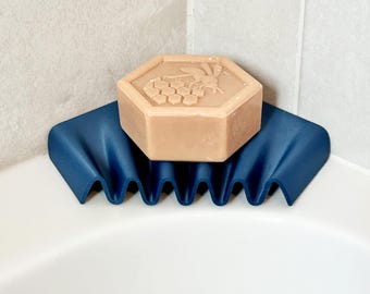 May include: A hexagonal bar of soap with a bee and honeycomb design rests on a dark blue soap dish. The soap dish has a wavy design and is placed in the corner of a white bathtub.