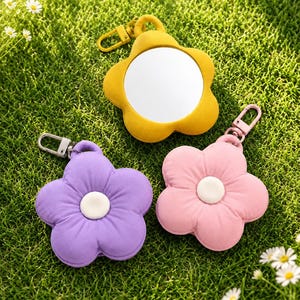 May include: Three flower-shaped accessories with keychains. One is yellow with a mirror, and the other two are purple and pink with white centers. The accessories are on a bed of green grass.
