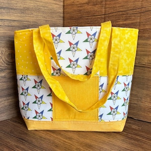 Peut inclure: Un sac fourre-tout jaune avec un panneau blanc orné d'un motif d'étoiles répétées en rouge, bleu, vert et jaune. Le sac a des poignées jaunes et une poche jaune. Le sac est en tissu.