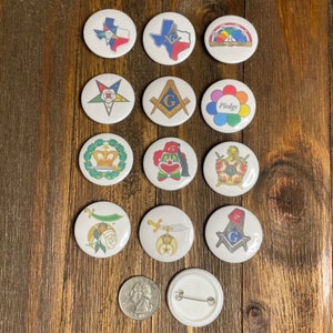 Puede incluir: Doce botones redondos blancos con varios diseños coloridos. Los diseños incluyen un payaso, una corona, un arcoíris, una brújula, una estrella, una espada y una bandera de Texas. Los botones están dispuestos en tres filas de cuatro.