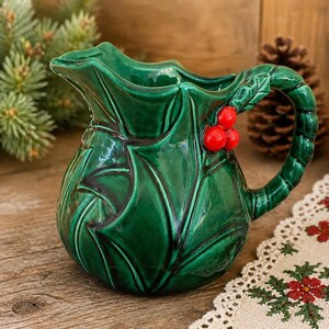 May include: A green ceramic pitcher with a holly leaf design. The pitcher has a handle and is adorned with three red berries. The pitcher is a festive holiday decoration.