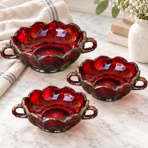 May include: Three ruby red glass bowls with two handles each, featuring a scalloped rim and a pressed starburst pattern on the base. The bowls are translucent and have a reflective surface, showcasing a deep red color throughout the glass structure.