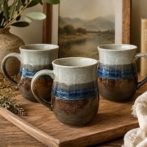 May include: Three ceramic mugs with a rounded shape and a handle on the side. Each mug features a multi-toned glaze with a light cream top, a vibrant blue band in the middle, and a dark brown base with a speckled, textured finish throughout.