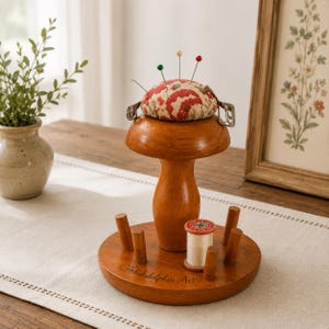 May include: A wooden mushroom-shaped sewing organizer with a round base featuring the text Philadelphia Art. The top is a fabric pincushion with red floral patterns and three pins. A small spool of white thread sits on the base among wooden pegs.