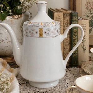 May include: A white porcelain teapot featuring a tapered body, a curved handle, and a matching lid. The upper section is decorated with a repeating pattern of gold and grey floral motifs and geometric accents. The teapot has a smooth, glossy finish.