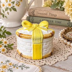 May include: A round ceramic trinket box designed to look like a gift basket. The white base features a textured woven pattern with brown bamboo-style trim. A bright yellow ceramic ribbon and bow are molded onto the lid, completing the gift look.