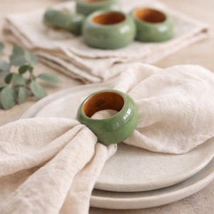 May include: A green ceramic napkin ring with a wooden interior sits atop a folded white linen napkin. Several other napkin rings are visible in the background. The napkin rings are part of a dining set.