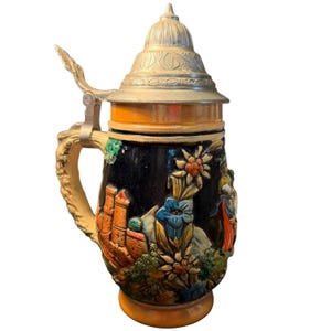 May include: A decorative beer stein with a hinged metal lid. The ceramic body is black with colorful relief images of flowers, a castle, and figures. The handle is beige and the base is orange.