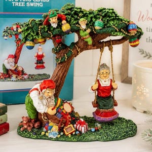 May include: A festive Christmas figurine depicting a tree with Santa and Mrs. Claus. Santa is shown with a sack of toys, while Mrs. Claus is on a swing. The tree is adorned with baubles and lights. The base is green with a grassy texture.