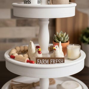 May include: A three-tiered white serving tray with farmhouse decor. The tray holds ceramic chickens, a small rolling pin, wooden beads, a succulent in a terracotta pot, and a candle. A wooden sign reads "FARM FRESH."