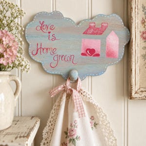 May include: A cloud-shaped wooden wall hook painted in light blue with a pink house graphic and the text "Love is Home grown" in pink script. A white towel with a pink gingham ribbon and lace trim hangs from the central wooden peg.