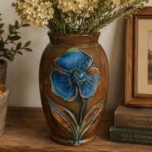 May include: A brown ceramic vase featuring a raised, hand-painted blue flower design with green leaves on the front. The vase has a smooth, glossy finish and a tapered neck, holding a bouquet of small, light-colored flowers inside the opening.