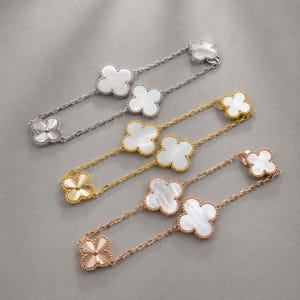 Op de afbeelding: Vier kettingen met klavervormige hangers in zilver, goud en roségoud. Elke ketting heeft vierbladige klaverhangers met witte parelmoer centra. De kettingen zijn delicaat, op een grijze ondergrond.