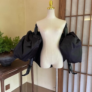 May include: A black, voluminous sleeve top displayed on a mannequin. The sleeves are large and puffy, creating a dramatic silhouette. The mannequin is on a metal stand, and the background is a neutral color.