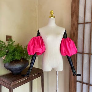 May include: A dress form with black arms and a gold-colored top. The form is wearing bright pink, voluminous sleeves. The background is a neutral color with a wooden door and a table with a plant.