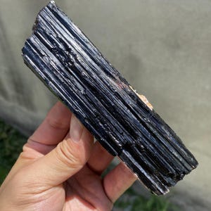 Puede incluir: Un cristal de turmalina negra en bruto con una textura estriada. El cristal es de un negro profundo y brillante con sutiles variaciones de tono. La superficie tiene un aspecto natural y sin pulir. El cristal se sostiene en una mano.