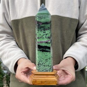 Può includere: Obelisco in cristallo di zoisite rettangolare e lucido. La pietra presenta un colore di base verde brillante con inclusioni nere e magenta. Montato su una piccola base quadrata in legno.
