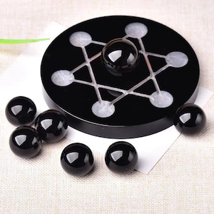 May include: A black, round, decorative stand with a white star-like design and a black sphere in the center. Six additional black spheres are scattered around the stand, creating a balanced composition. The objects are on a white surface.
