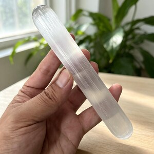 May include: A cylindrical selenite crystal wand with a smooth, polished surface and a translucent white appearance. The stone features natural vertical striations and a rounded tip, showcasing a fibrous texture that catches the light.