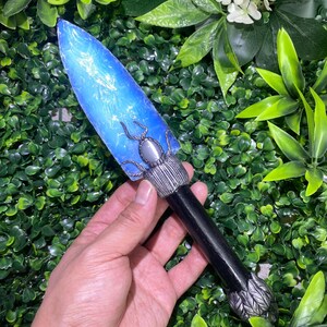 Puede incluir: Una daga decorativa con una hoja azul translúcida y un diseño de insecto plateado. El mango es negro con detalles plateados. La daga mide aproximadamente 20 cm de largo.