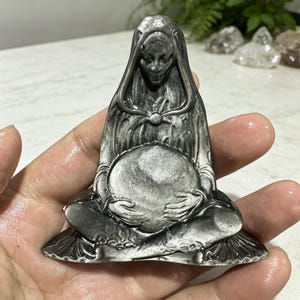 May include: A silver obsidian carving of a hooded figure in a seated position, holding a large, smooth, circular sphere in their lap. The figure features intricate folds in the clothing and a detailed face, with a metallic, reflective sheen throughout.