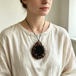 May include: A dark brown pendant necklace featuring a large, smooth black oval center stone. The frame is crafted with intricate clay-like swirls and claw-like prongs, accented by small, embedded amber and red gemstones on a black cord.