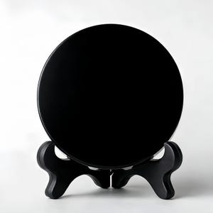 May include: A glossy black, round decorative plate displayed on a black wooden stand. The plate is smooth and reflective, and the stand has a three-legged design. The background is plain white.