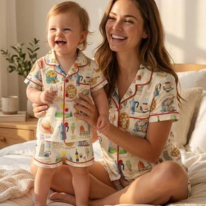 K&ouml;nnte beinhalten: Passende Pyjama-Sets mit cremefarbenem Hintergrund und einem bunten Lebensmittel- und Getr&auml;nkemuster. Die Sets bestehen aus einem kurz&auml;rmeligen Oberteil und Shorts, rot eingefasst. Eine Frau und ein Kind tragen die Pyjamas und l&auml;cheln.