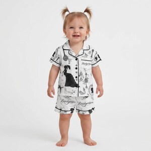 Op de afbeelding: Witte baby pyjamaset met een zwart-wit patroon met silhouetten en het woord "Bridgerton". De top met korte mouwen en de shorts hebben een zwarte bies. Het kind draagt de pyjama.