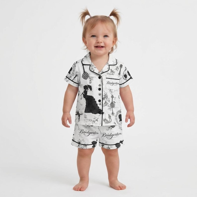 Op de afbeelding: Witte baby pyjamaset met een zwart-wit patroon met silhouetten en het woord "Bridgerton". De top met korte mouwen en de shorts hebben een zwarte bies. Het kind draagt de pyjama.