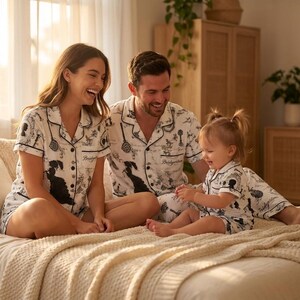 Op de afbeelding: Een gezin draagt bijpassende witte pyjamasets met zwarte illustraties. De sets bevatten shirts met korte mouwen en shorts. Het woord "Bridgerton" staat op de shirts. Het gezin zit op een bed.