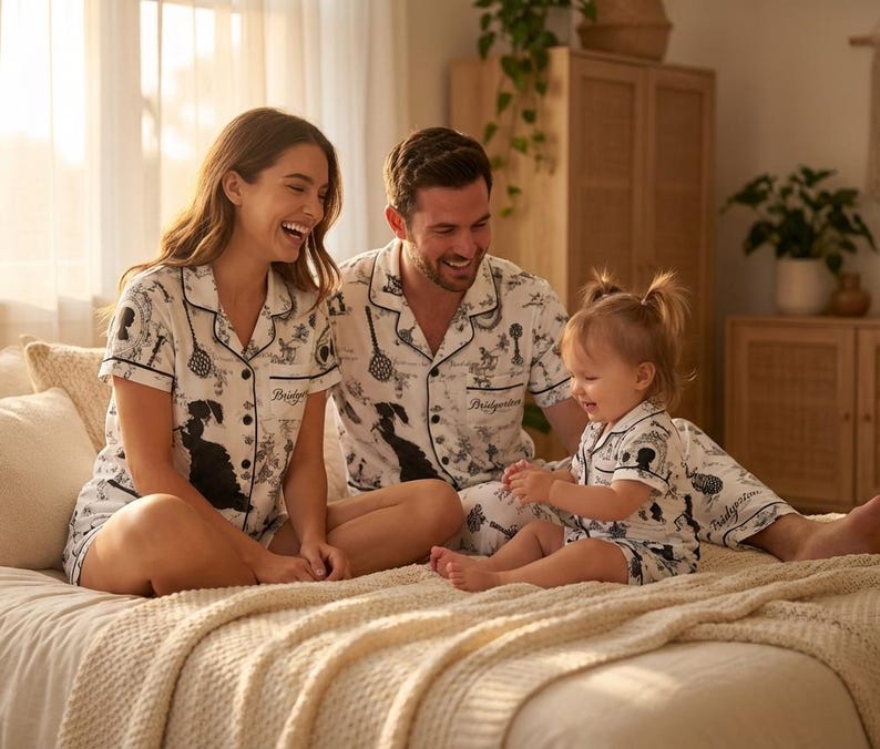 Op de afbeelding: Een gezin draagt bijpassende witte pyjamasets met zwarte illustraties. De sets bevatten shirts met korte mouwen en shorts. Het woord "Bridgerton" staat op de shirts. Het gezin zit op een bed.
