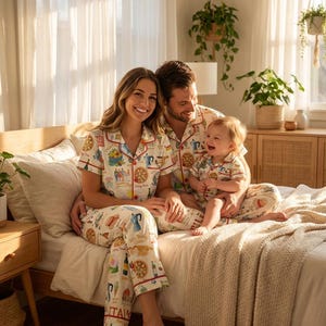 K&ouml;nnte beinhalten: Passende Familien-Pyjama-Sets mit italienischem Thema. Die wei&szlig;en Pyjamas zeigen farbenfrohe Illustrationen italienischer Speisen und Symbole. Die Sets umfassen kurz&auml;rmelige Hemden und Hosen mit rotem Besatz. Eine dreik&ouml;pfige Familie tr&auml;gt die Pyjamas auf einem Bett.