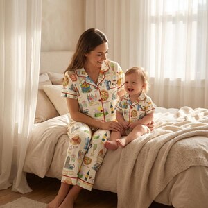K&ouml;nnte beinhalten: Passende Pyjama-Sets. Die cremefarbenen Pyjamas haben einen bunten Aufdruck mit roter Bord&uuml;re. Der Erwachsene tr&auml;gt ein lang&auml;rmliges Oberteil und eine Hose, w&auml;hrend das Kind ein kurz&auml;rmliges Oberteil und Shorts tr&auml;gt. Beide sitzen auf einem Bett.