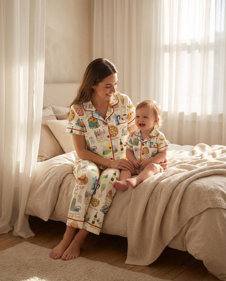 K&ouml;nnte beinhalten: Passende Pyjama-Sets. Die cremefarbenen Pyjamas haben einen bunten Aufdruck mit roter Bord&uuml;re. Der Erwachsene tr&auml;gt ein lang&auml;rmliges Oberteil und eine Hose, w&auml;hrend das Kind ein kurz&auml;rmliges Oberteil und Shorts tr&auml;gt. Beide sitzen auf einem Bett.