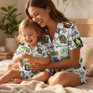 May include: Matching white pajama sets with a green and purple graphic print. The sets feature a button-down shirt and shorts. The print includes images of characters and the word "Ghouls". The mother and child are smiling.
