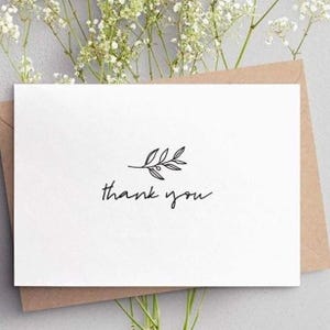 K&ouml;nnte beinhalten: Eine wei&szlig;e Karte mit dem handgeschriebenen Schriftzug "thank you" und einer kleinen Bl&auml;tterillustration. Die Karte liegt auf einem braunen Umschlag, mit zarten wei&szlig;en Blumen im Hintergrund.