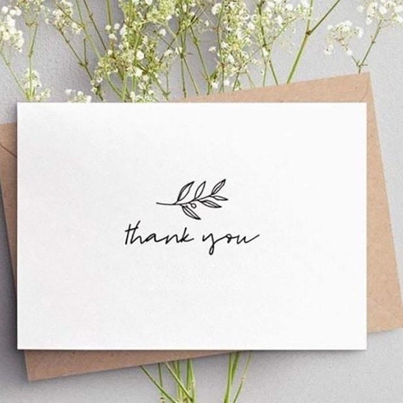 K&ouml;nnte beinhalten: Eine wei&szlig;e Karte mit dem handgeschriebenen Schriftzug "thank you" und einer kleinen Bl&auml;tterillustration. Die Karte liegt auf einem braunen Umschlag, mit zarten wei&szlig;en Blumen im Hintergrund.
