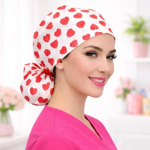 May include: A white scrub cap with a pattern of red hearts. The cap is tied in the back and is worn by a person wearing a pink scrub top. The background is blurred, with hints of plants and other objects.