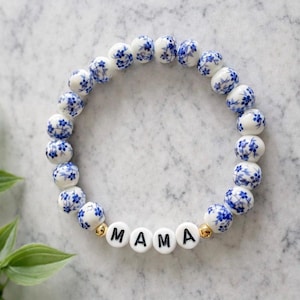 May include: A beaded bracelet featuring white and blue floral patterned beads. The bracelet includes white letter beads spelling "MAMA" and gold accent beads. The bracelet is a stylish accessory.