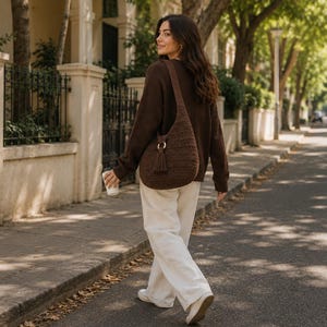 May include: A dark brown crochet hobo bag featuring a wide shoulder strap, a circular wooden ring accent, and a matching tassel detail. The bag is worn over a dark brown long-sleeved jumper paired with loose-fitting white trousers and white shoes.