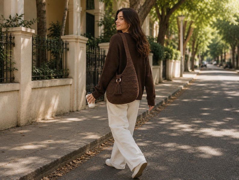 May include: A dark brown crochet hobo bag featuring a wide shoulder strap, a circular wooden ring accent, and a matching tassel detail. The bag is worn over a dark brown long-sleeved jumper paired with loose-fitting white trousers and white shoes.