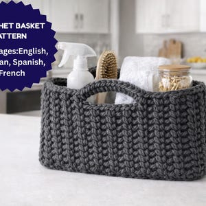 Peut inclure: Un panier en crochet gris foncé avec deux poignées, rempli d'un flacon pulvérisateur, d'une brosse, d'une serviette blanche et d'un bocal. Le panier est posé sur une surface blanche. Le texte sur un badge bleu indique "CROCHET BASKET PATTERN" et répertorie les langues.
